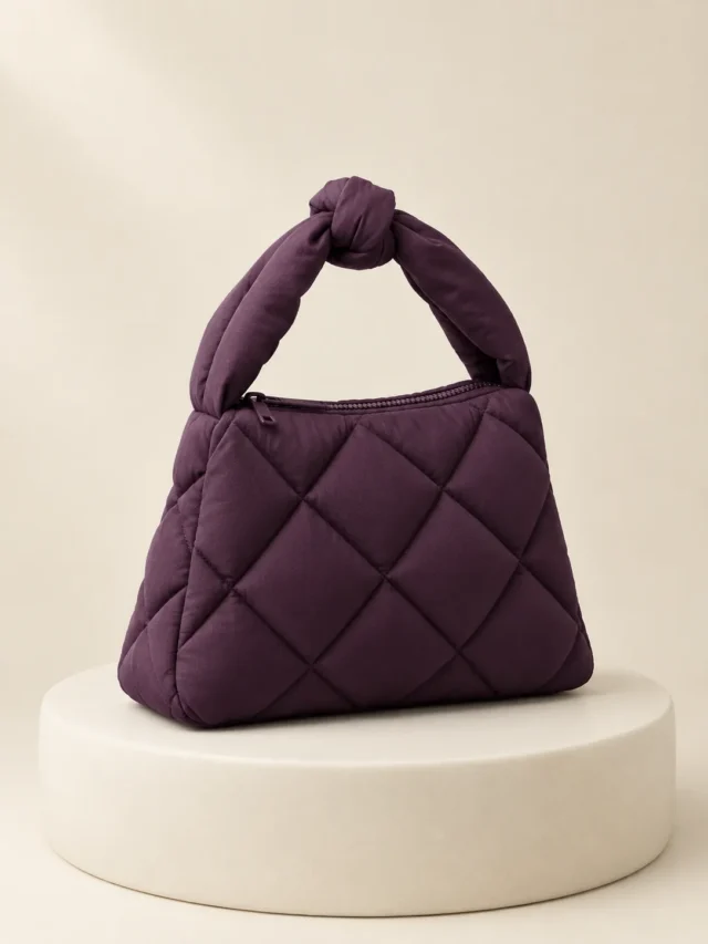 Knot Handle Puff in Plum, front view on a cream plinth